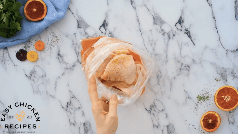 A person is holding a chicken in a plastic bag while preparing an orange chicken recipe.