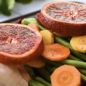 Blood orange slices sit atop a baking sheet filled with veggies and chicken thighs.