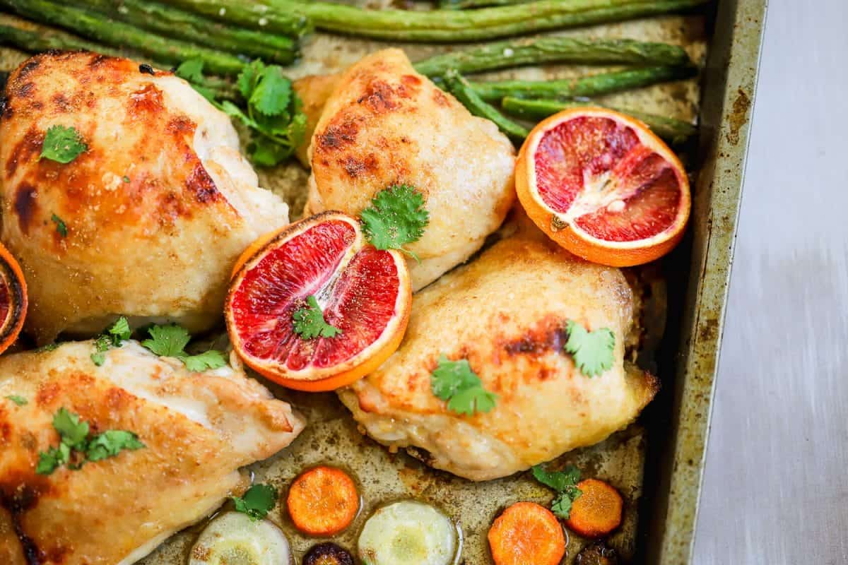 Blood orange chicken thighs with asparagus on a baking sheet.