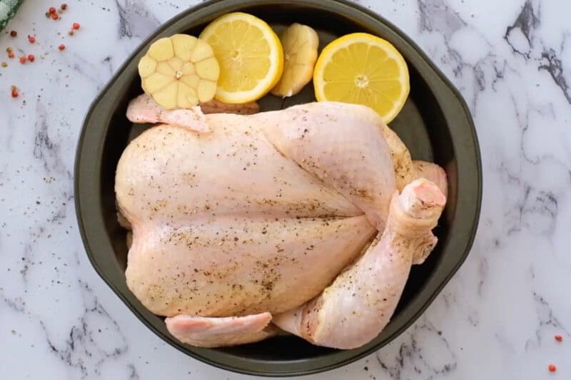 a raw whole seasoned chicken in a skillet with lemon slices and halved garlic.