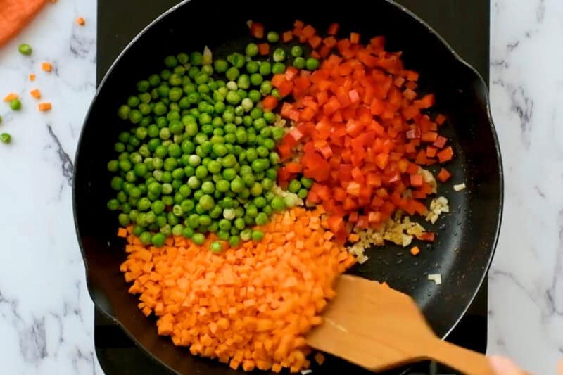 diced carrots, red bell peppers, and peas in a pan with onion.