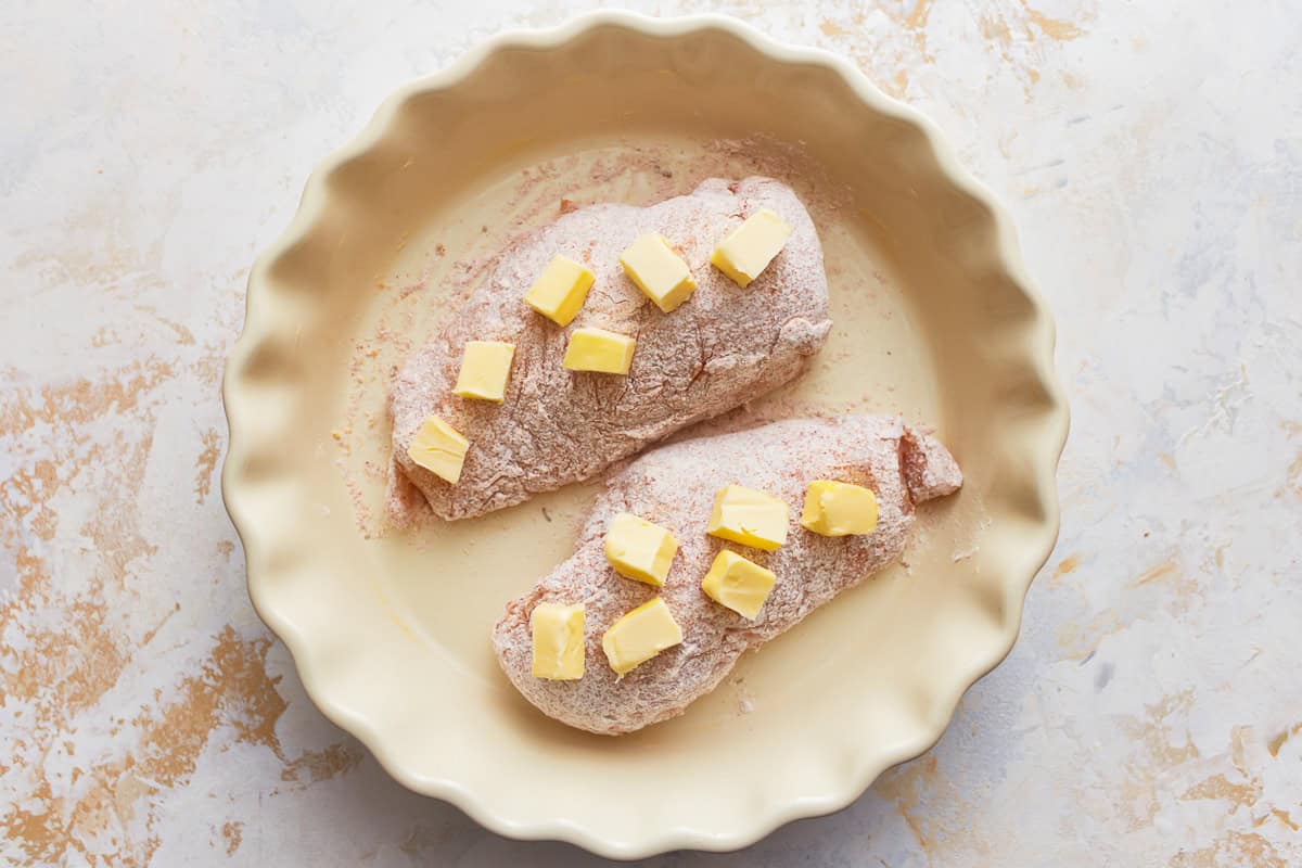 cubes of butter arranged over 2 flour-dredged chicken breasts in a baking dish.