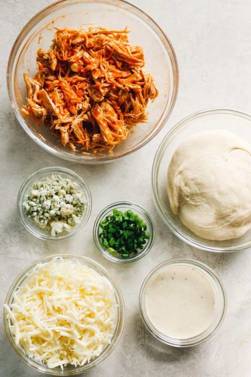 Buffalo chicken pizza ingredients displayed in bowls on a table.