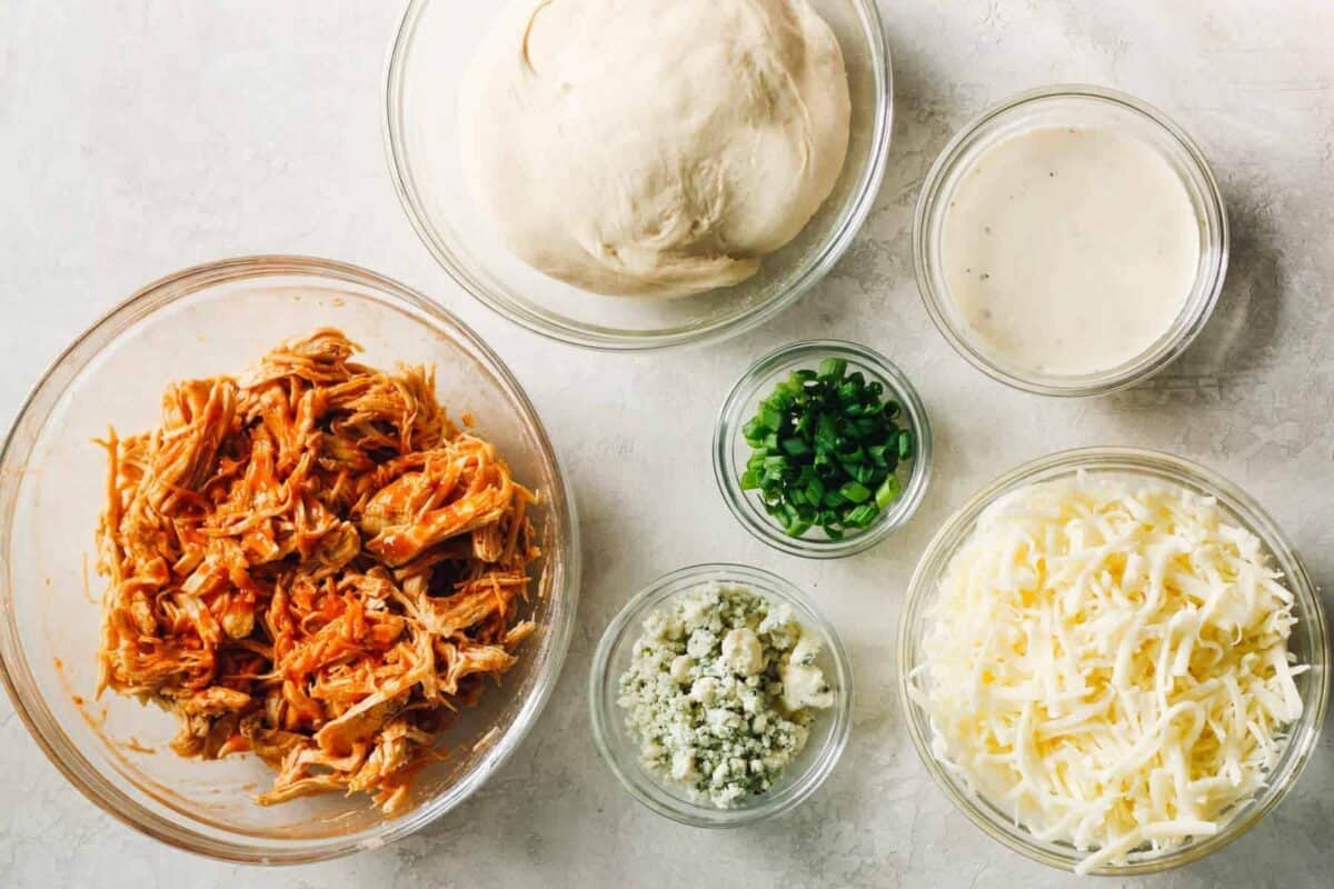 Buffalo chicken pizza ingredients displayed in bowls on a table.