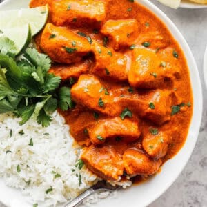 overhead view of a serving of butter chicken in a white bowl with cilantro, rice, and lime wedges.