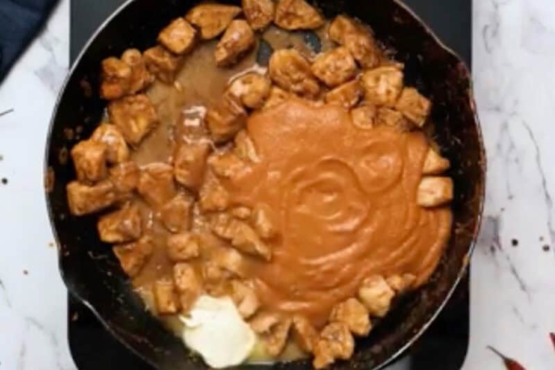adding butter and butter chicken sauce to cooked chicken pieces in a skillet.