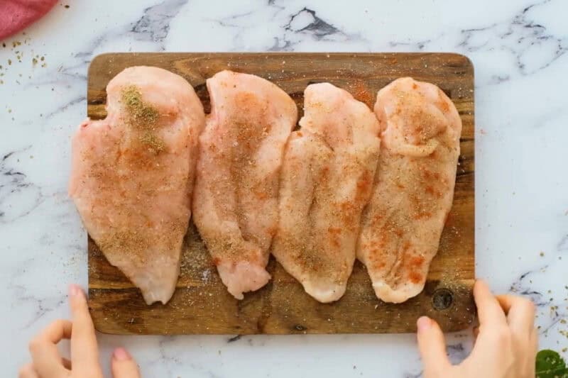 four seasoned raw chicken breasts on a wooden cutting board.