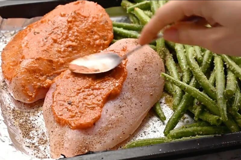 spreading sun dried tomato pesto over two raw seasoned chicken breasts on a baking sheet.