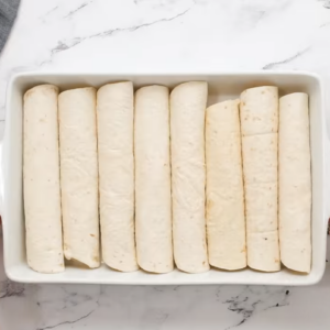 Tortillas are rolled with filling and lined up in a casserole dish.