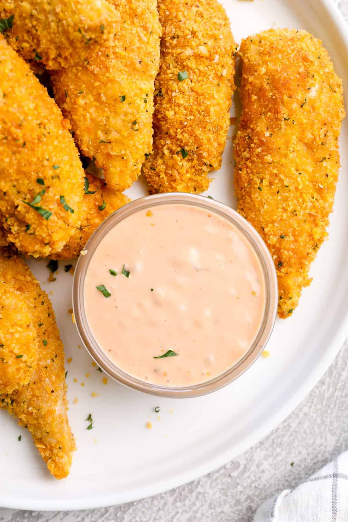 Cornflake-crusted chicken fingers on a plate with a small bowl of dipping sauce.