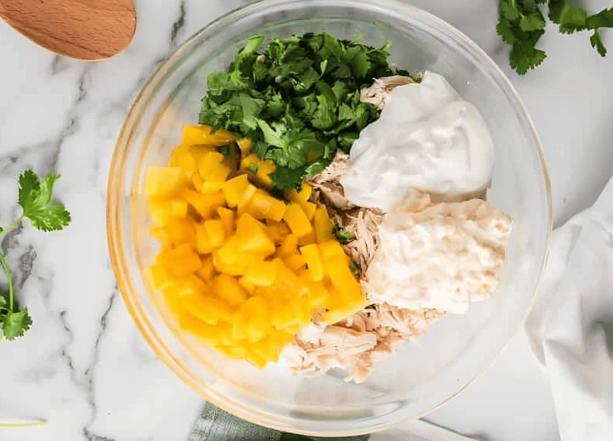 yogurt, mayo, mango, and cilantro over shredded cooked chicken in a glass bowl.