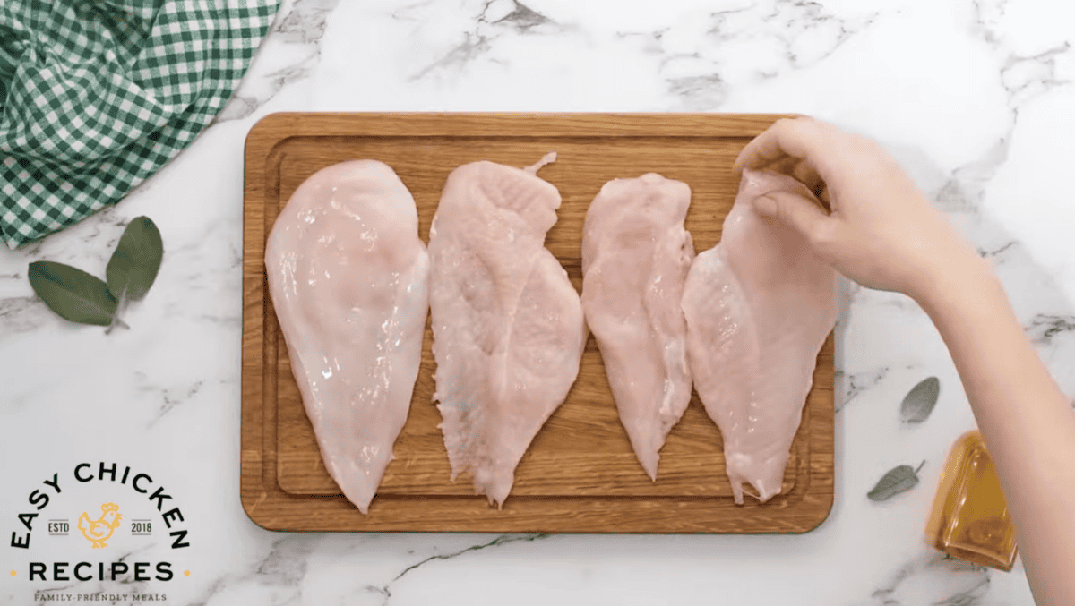 Four flattened chicken breasts are on a wooden cutting board. 