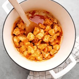 cubed chicken in red sauce in a dutch oven with a wooden spoon.