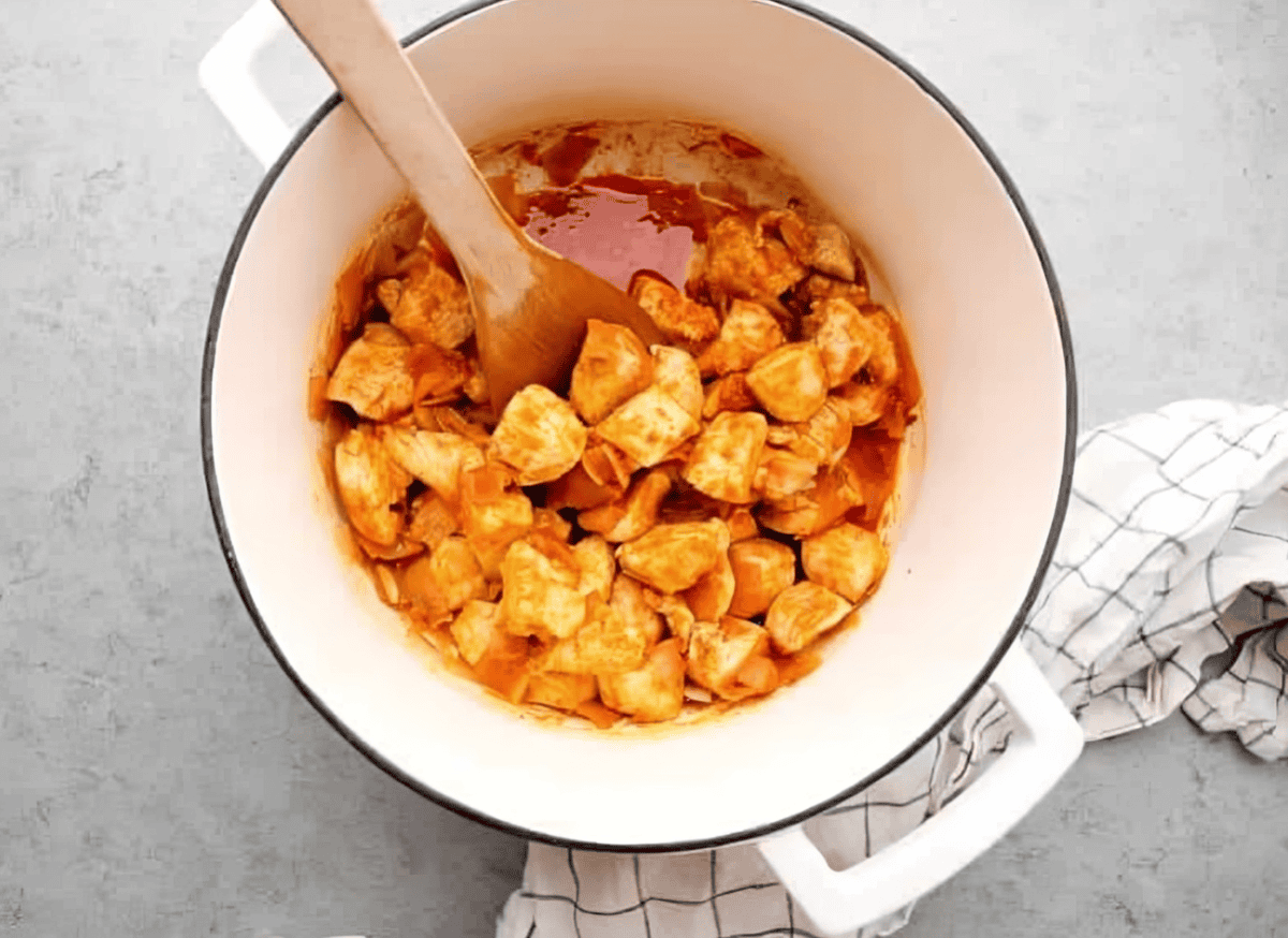 cubed chicken in red sauce in a dutch oven with a wooden spoon.