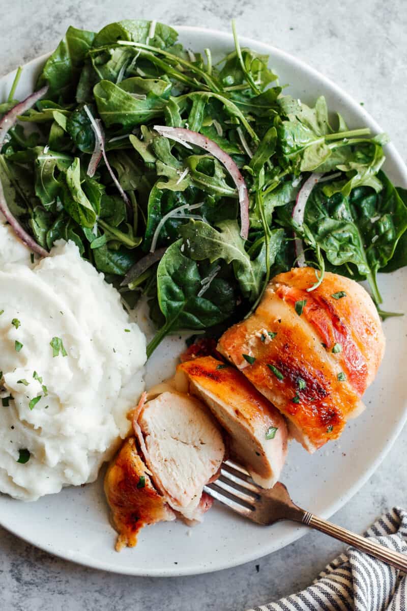 sliced bacon wrapped chicken next to salad and mashed potatoes