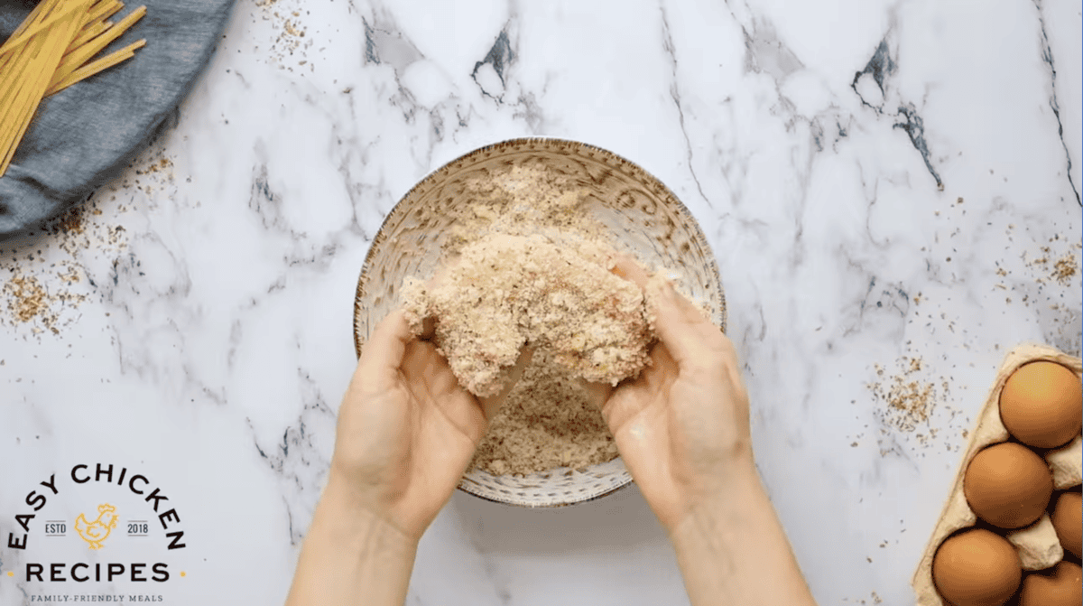 holding a breaded raw chicken thigh over a bowl of breadcrumbs.