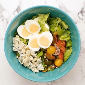 lettuce topped with halved cherry tomatoes, halved hard boiled eggs, and blue cheese in a blue bowl.