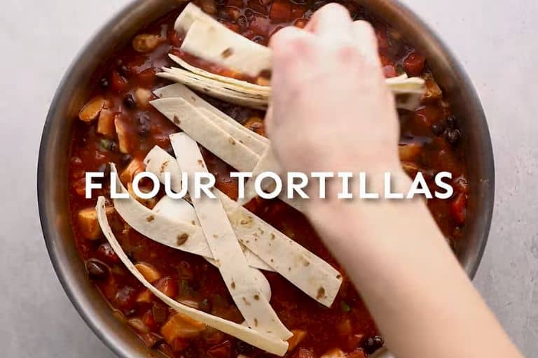 A hand adding flour tortilla strips to chicken enchilada casserole in a skillet.