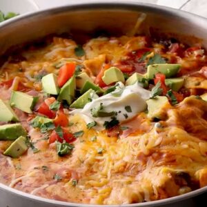 Chicken enchilada casserole topped with avocados, tomatoes, and cilantro in a skillet.