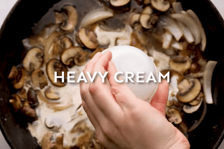 A hand pouring heavy cream into a skillet with mushrooms and marsala wine.