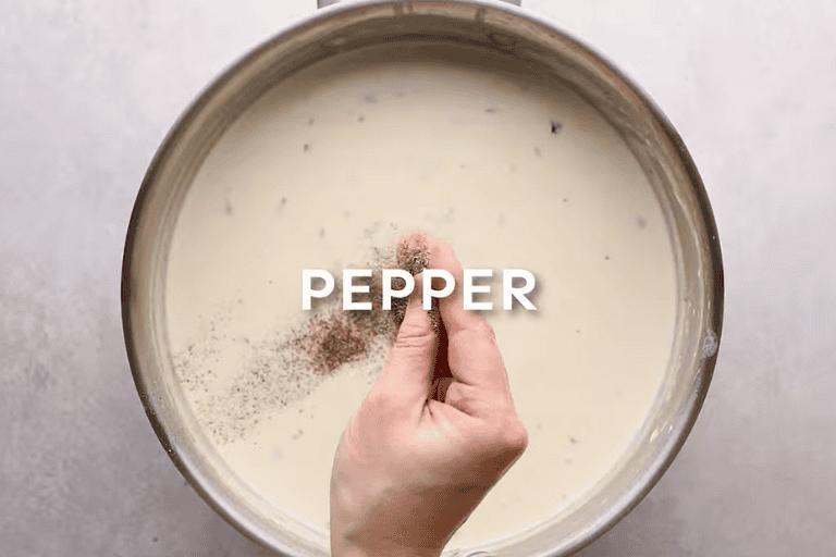 A hand adding ground pepper to a pot of alfredo sauce.