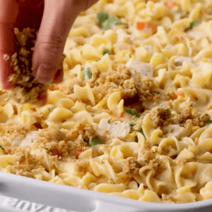 A hand sprinkling breadcrumbs onto a chicken noodle casserole.
