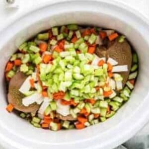 Raw chicken breasts topped with diced carrots, onions, and celery in a crockpot.