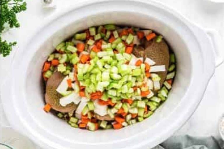 Raw chicken breasts topped with diced carrots, onions, and celery in a crockpot.