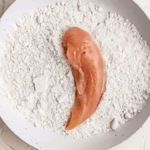 a raw chicken tender in a bowl of flour.