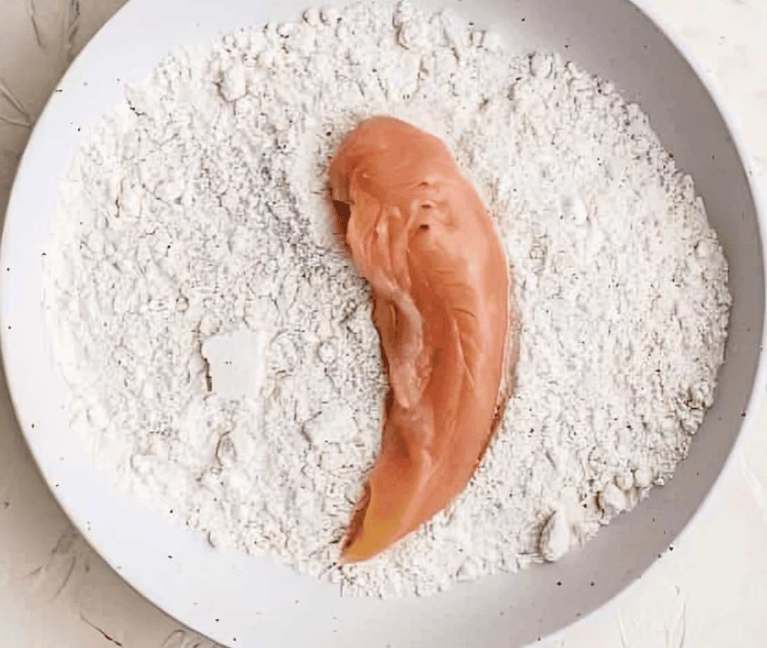 a raw chicken tender in a bowl of flour.