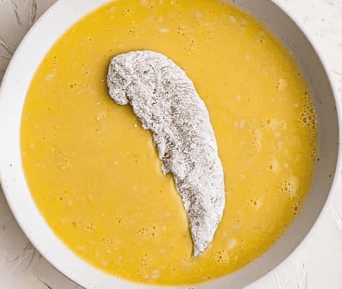 a raw flour-coated chicken tender in a bowl of raw eggs.