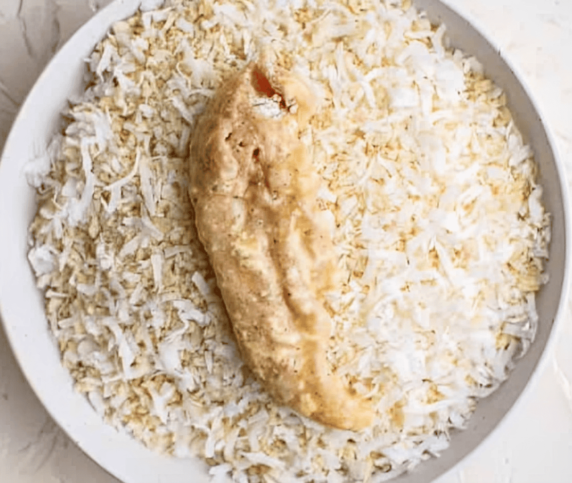 a raw, flour-and-egg-coated chicken tender in a bowl of coconut breadcrumbs.