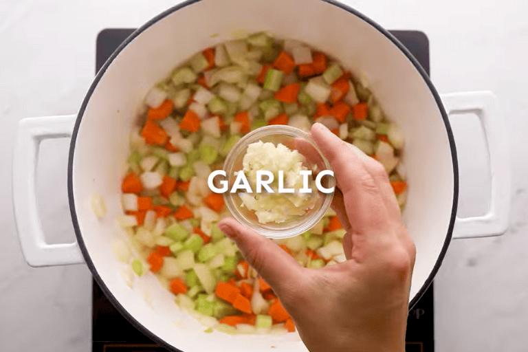 Minced garlic being added to a pot of carrots, celery, and onion.