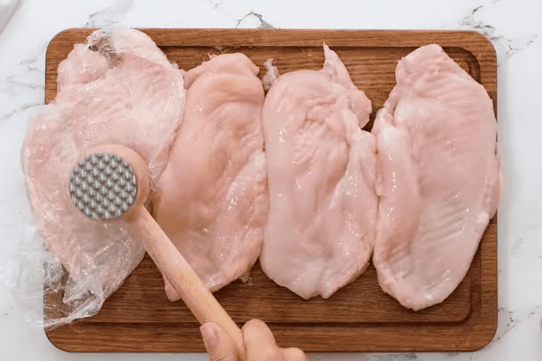 Four chicken breasts on a chopping board, being pounded with a meat mallet.