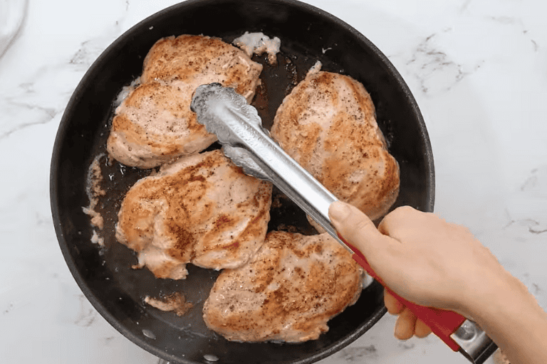 Four chicken breasts cooking a skillet, with a pair of tongs turning one over.