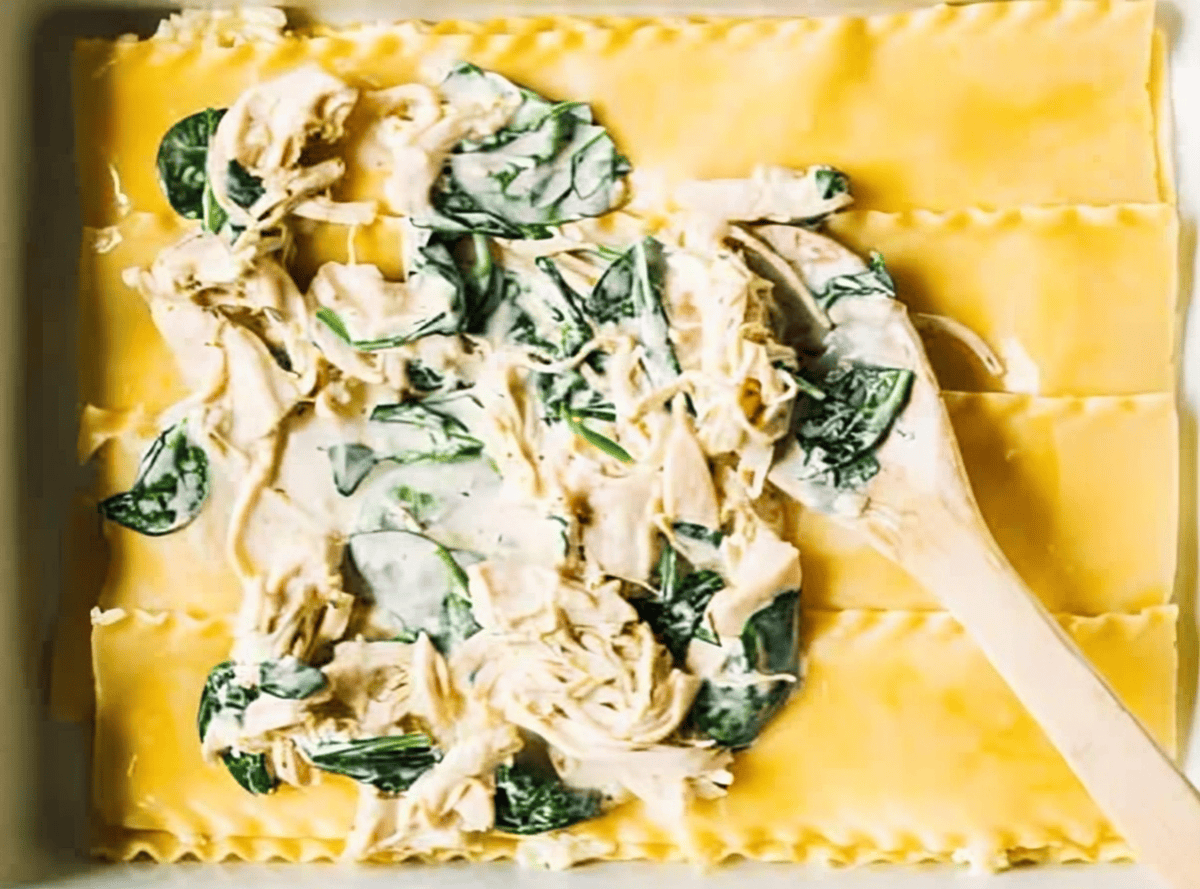 spreading chicken spinach filling over lasagna noodles with a wooden spoon.