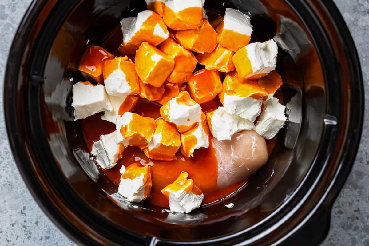 buffalo sauce drizzled over cubed cream cheese and raw chicken in a crockpot.