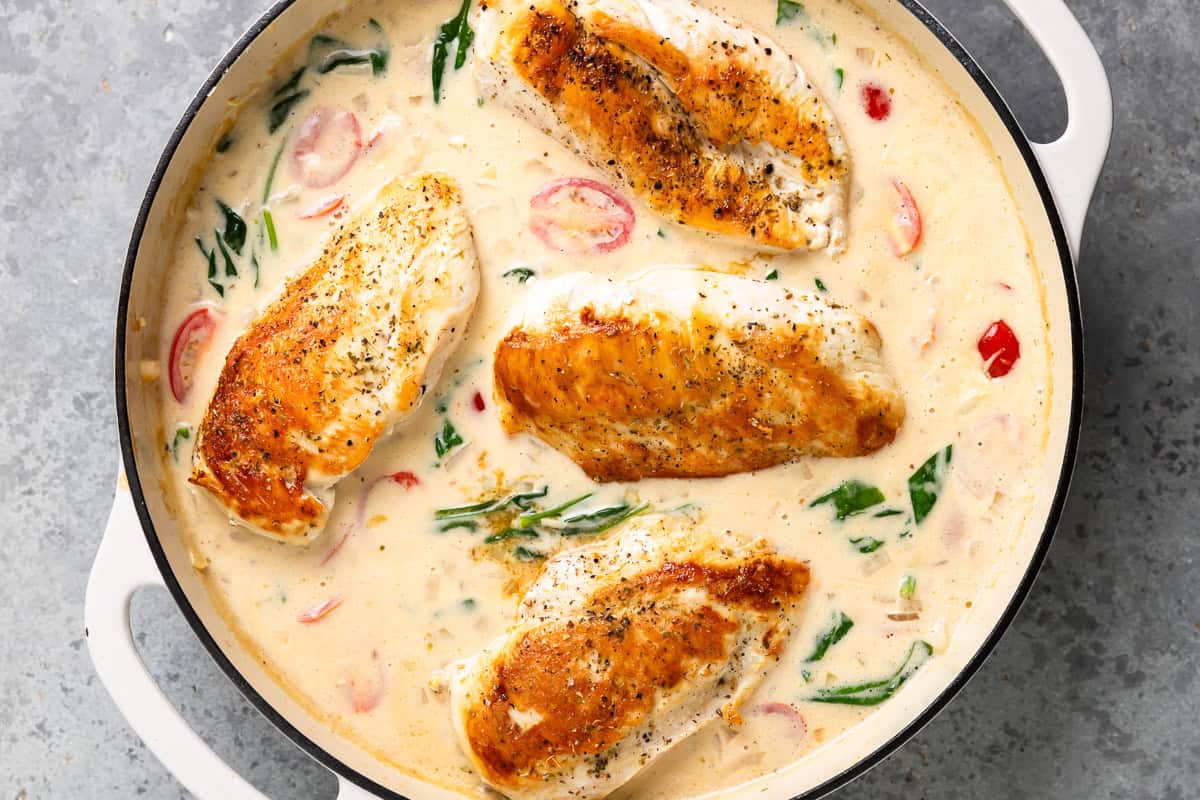 chicken added to cream sauce with spinach and tomatoes in a skillet.