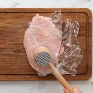 A chicken breast between plastic wrap being pounded with a meat mallet.