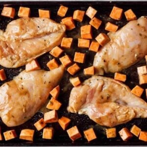 raw seasoned chicken breasts and cubed sweet potatoes on a sheet pan.