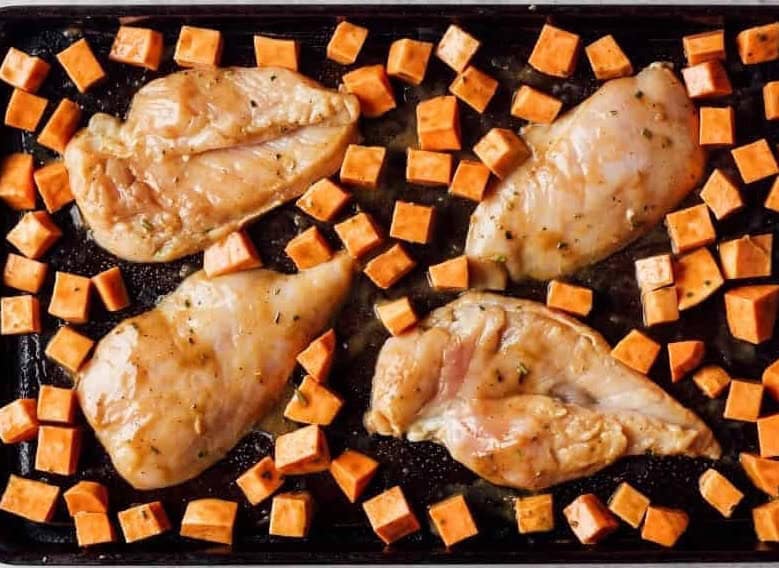 raw seasoned chicken breasts and cubed sweet potatoes on a sheet pan.