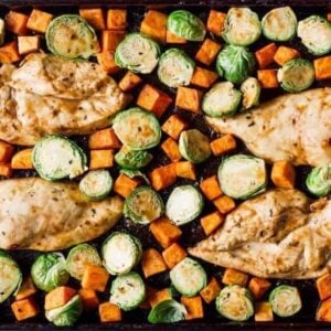 raw seasoned chicken breasts, cubed sweet potatoes, and halved brussels sprouts on a sheet pan.