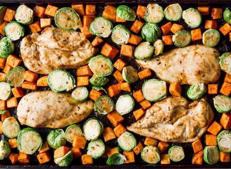 raw seasoned chicken breasts, cubed sweet potatoes, and halved brussels sprouts on a sheet pan.