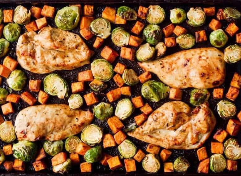 cooked chicken breasts, sweet potatoes, and brussels sprouts on a sheet pan.