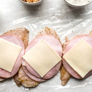 chicken breasts topped with ham and cheese on plastic wrap.