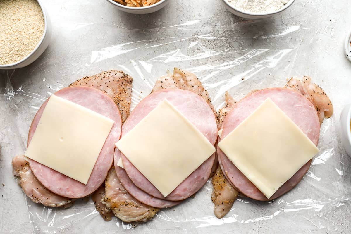 chicken breasts topped with ham and cheese on plastic wrap.