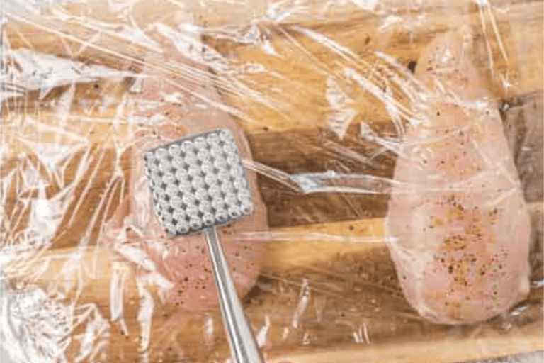 Seasoned chicken breasts covered in plastic wrap, being pounded by a meat mallet.