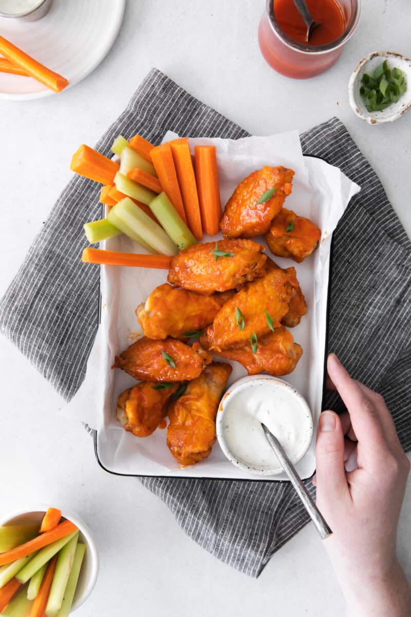 hand with a tray of honey buffalo chicken wings on a serving tray with sliced carrots and celery and dressing in a small bowl