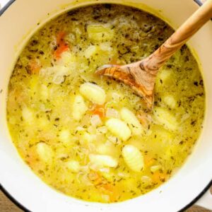 broth, gnocchi, spices, and veggies for chicken gnocchi soup in a white pot with a wood spoon