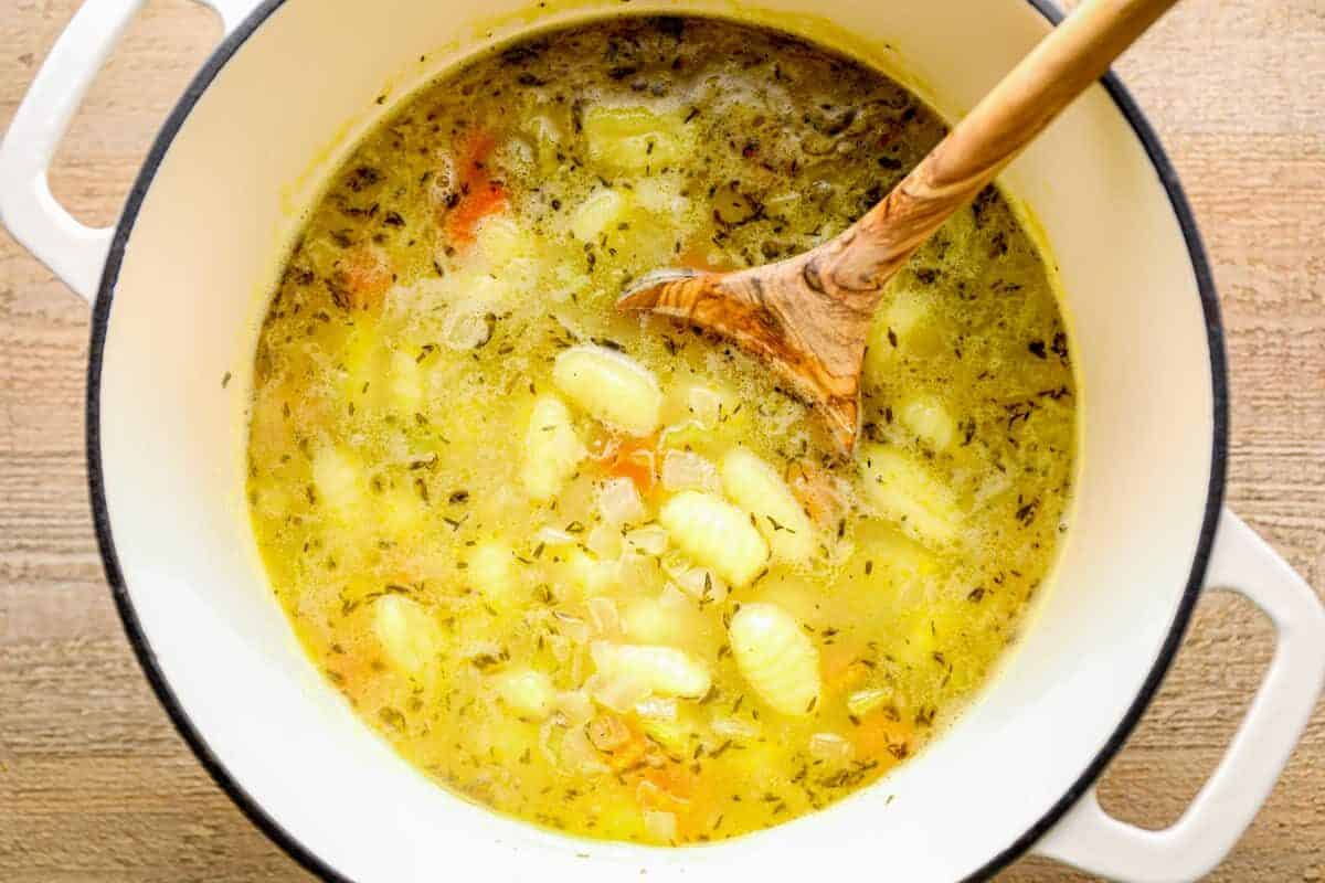 broth, gnocchi, spices, and veggies for chicken gnocchi soup in a white pot with a wood spoon